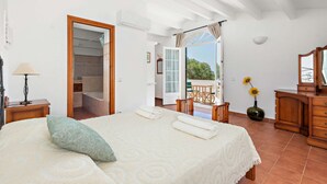 A bedroom with a bed, a nightstand, a lamp, a wooden dresser, a vase with sunflowers, and a view of a balcony and outside area. - New! Casa Nova de Biniparrell (Biniparrell)