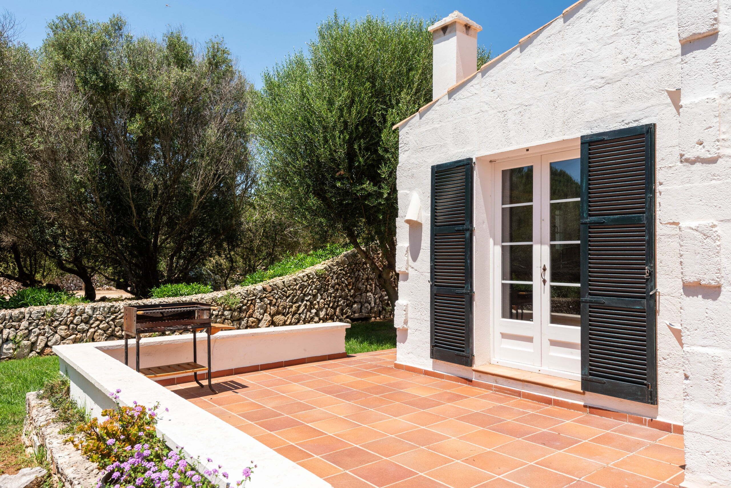 A white building with black shutters, a stone wall, and a tiled patio.