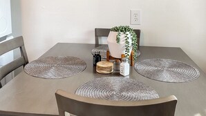 Un comedor con una mesa gris, dos individuales tejidos, una planta en maceta, saleros y pimenteros, y un cuadro abstracto decorativo en la pared.