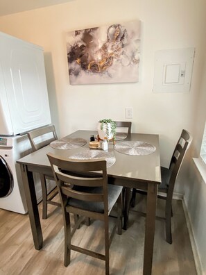 Een eetgedeelte met een tafel voor vier personen, omringd door stoelen, een aan de muur hangend schilderij en een wasmachine op de achtergrond.