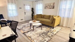 Una sala de estar con un sofá, una mesita de centro y una silla. Hay una puerta con la leyenda 'KEYS' y una ventana con cortinas.