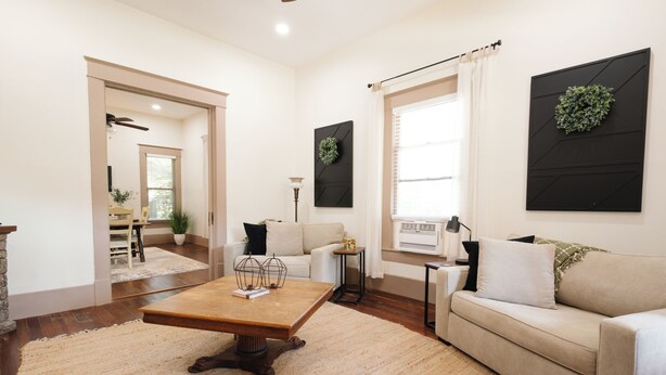 Un salon spacieux avec une table basse en bois, un canapé beige et un fauteuil blanc. On y trouve une grande fenêtre avec des rideaux, une œuvre d’art noire accrochée au mur et une plante en pot.