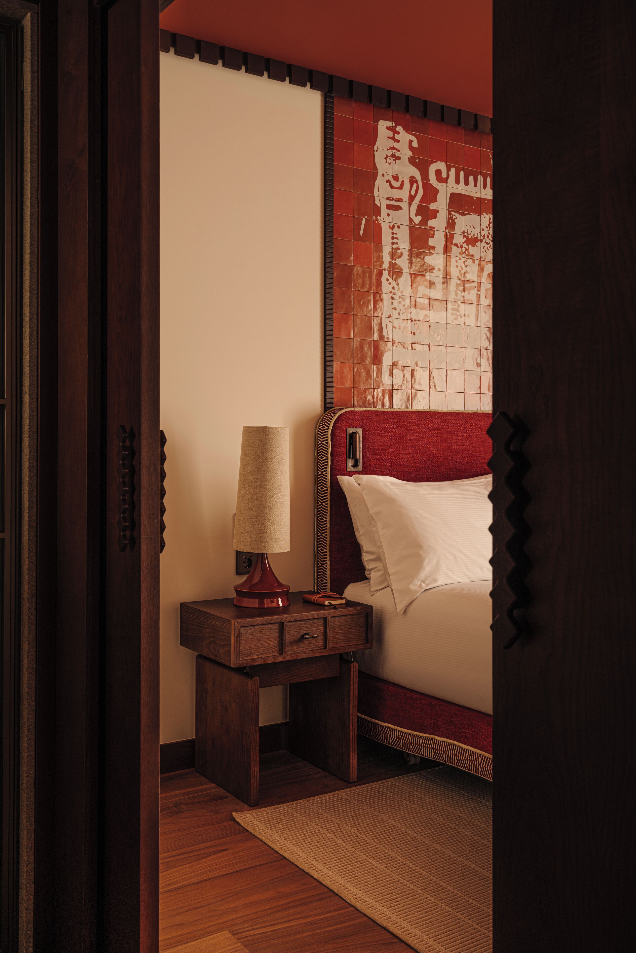 Un dormitorio con una mesita de noche de madera, una lámpara, una cama con cabecera roja y una alfombra estampada.