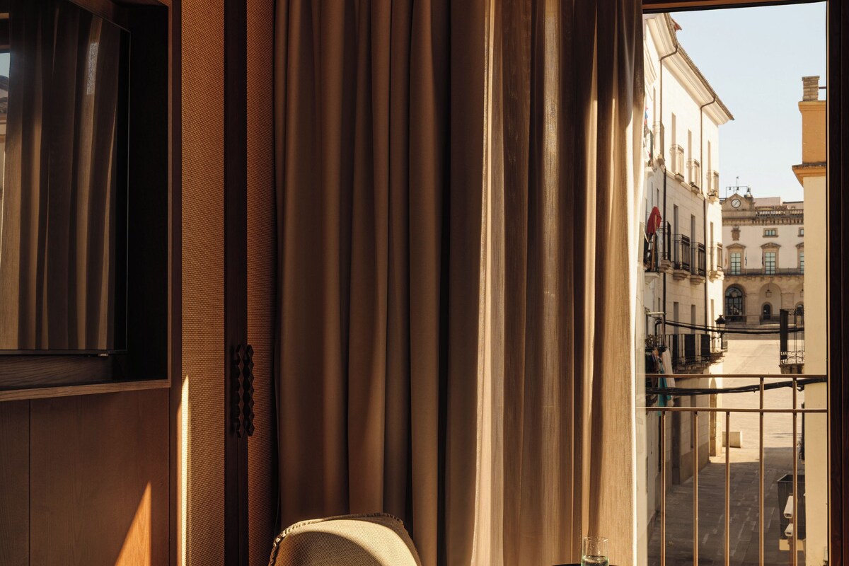 Quarto de hotel com cama, uma cadeira, uma mesa pequena e vista para a rua através de uma varanda.