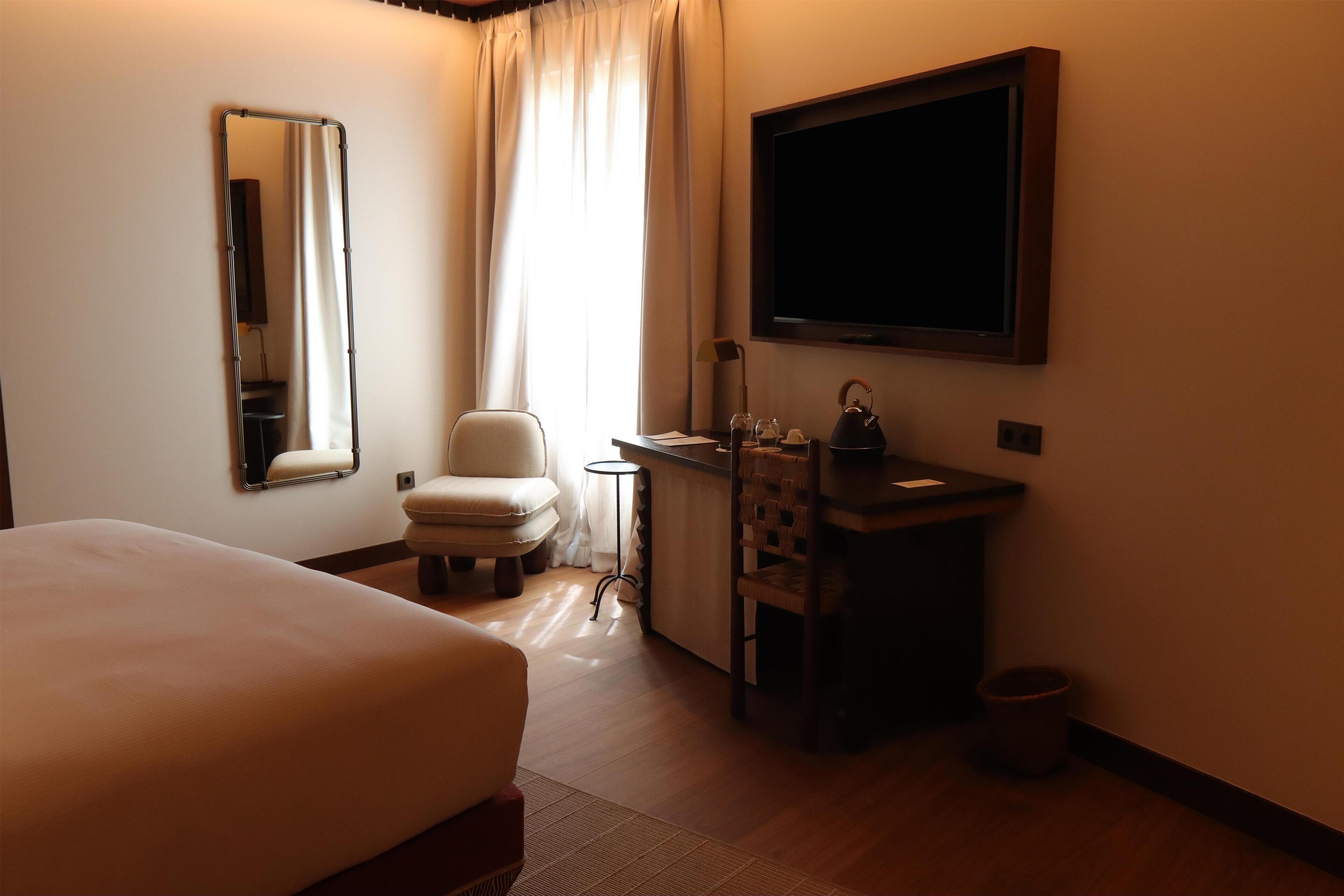 A hotel room with a bed, a chair, a desk with a sink, a mirror, and a TV.