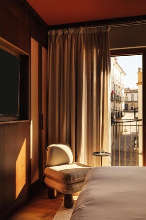 Ein Hotelzimmer mit einem großen Bett, einem Sessel, einem kleinen Tisch und Blick auf die Straße vom Balkon aus.