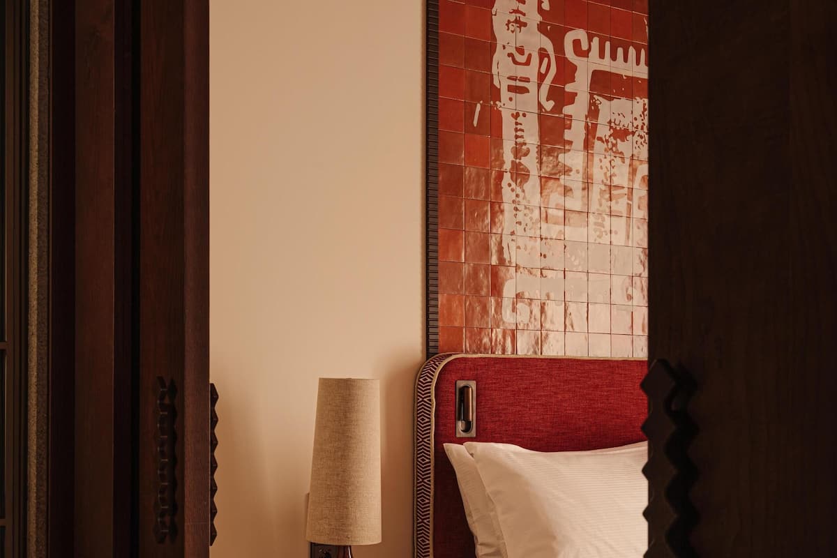 A bedroom with a wooden floor, a bedside table with a lamp, a bed with a red headboard, and a patterned wall.