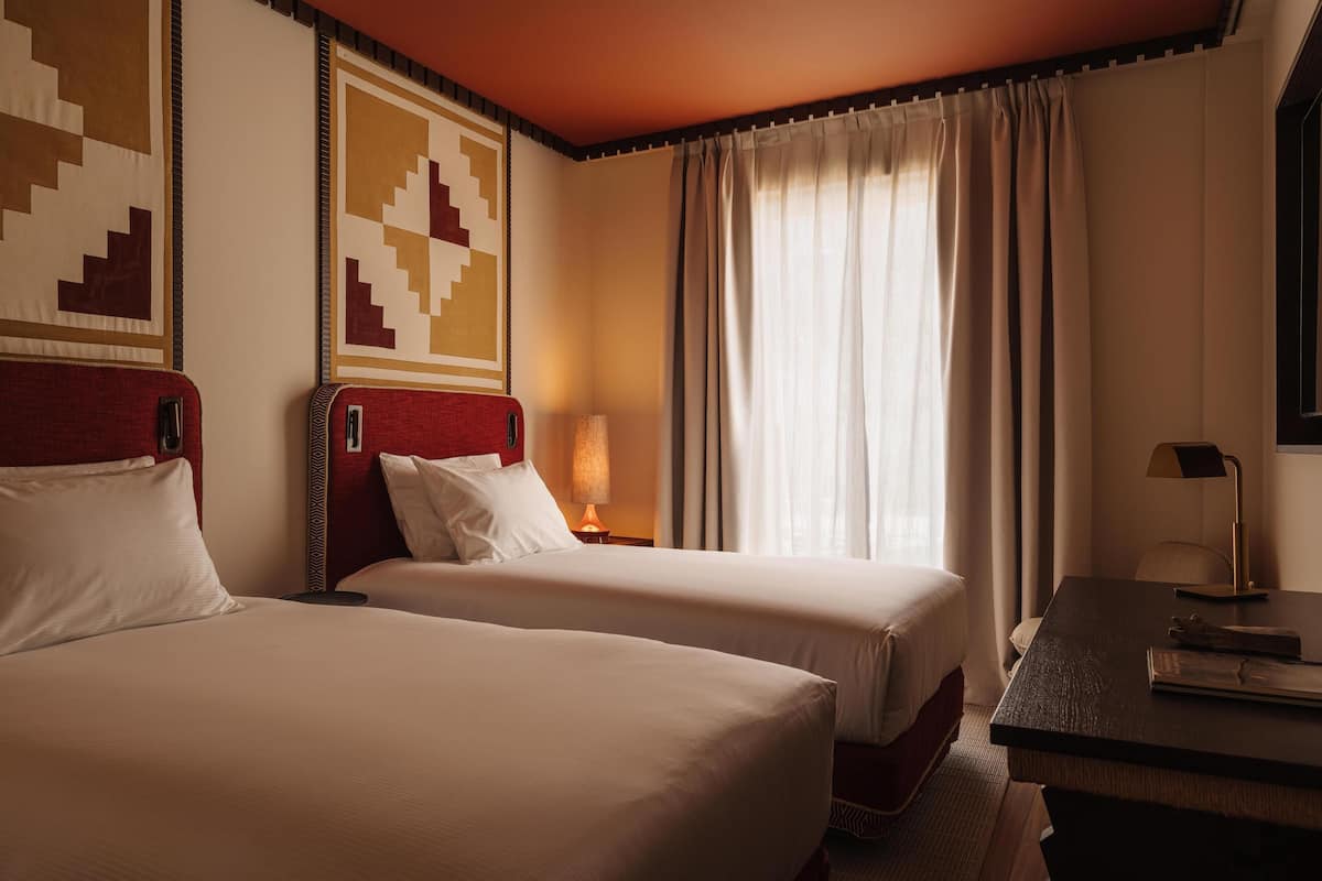 A hotel room with two beds, a desk, a lamp, and a window with curtains.
