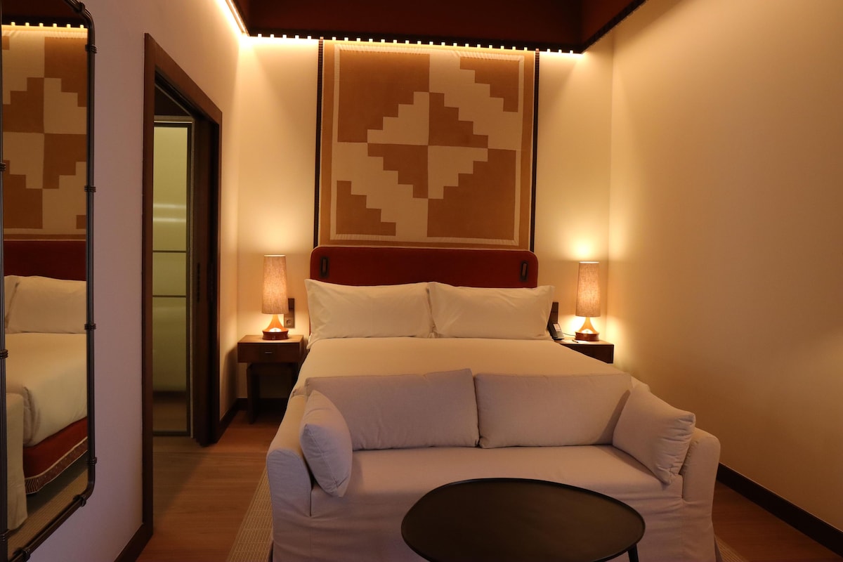 Quarto de hotel com uma cama, um sofá, duas mesinhas de cabeceira com abajures e um espelho grande.