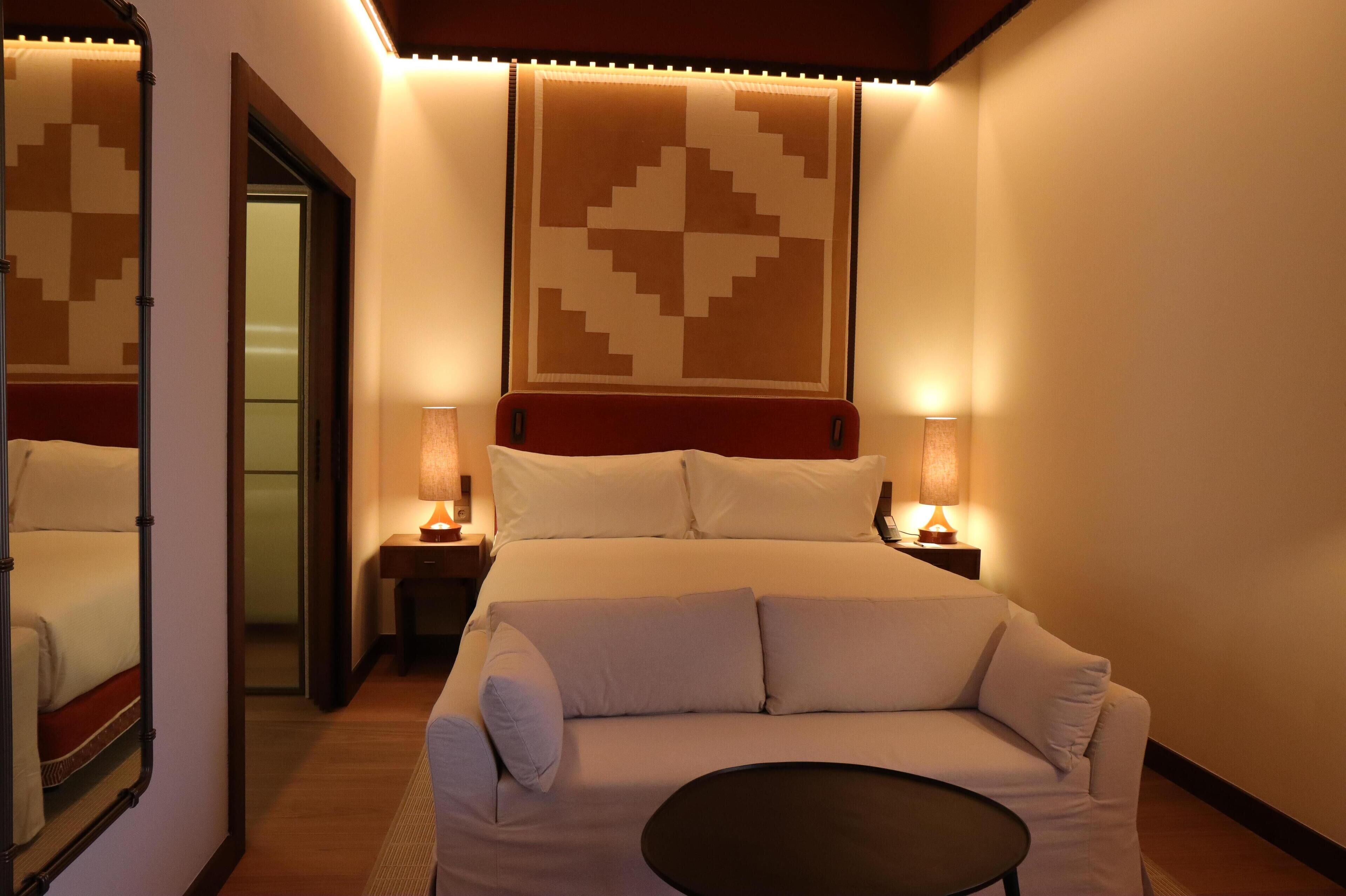 Habitación de hotel con cama, sofá, dos mesitas de noche con lámparas, un espejo y una pared con estampado geométrico.