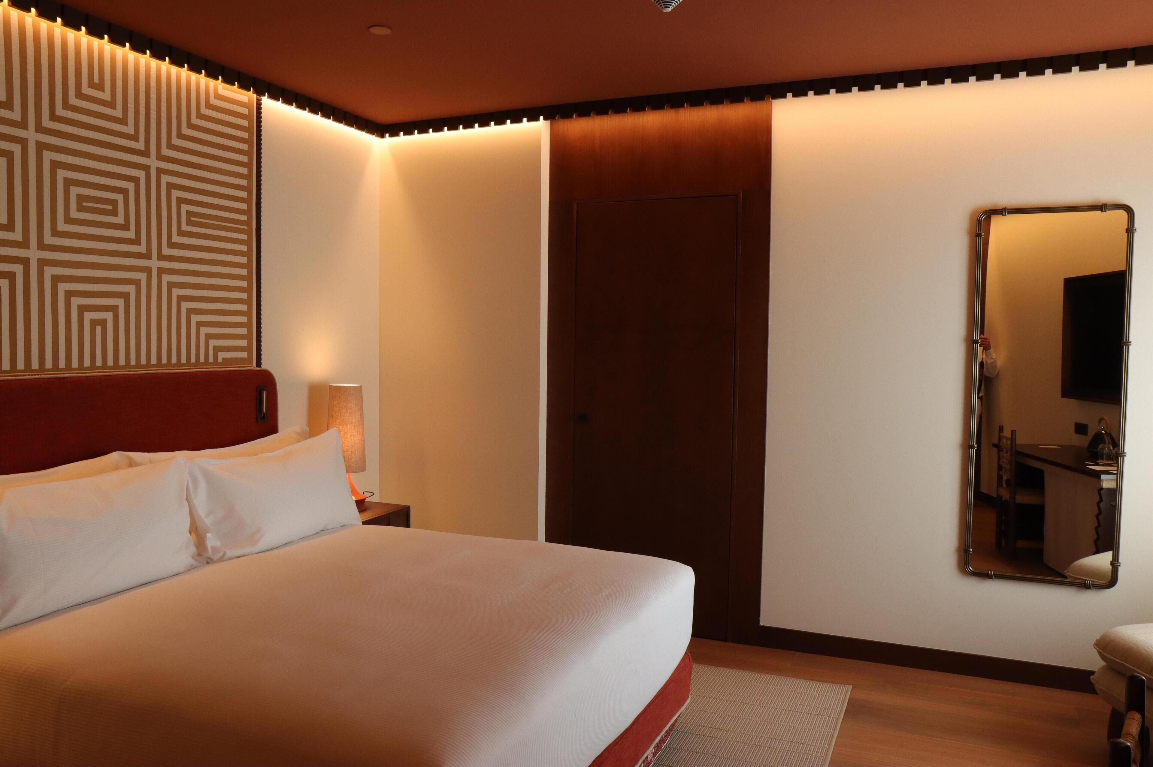 Una cama bien tendida con sábanas blancas, cabecera de madera, espejo montado en la pared y una puerta con panel de madera.