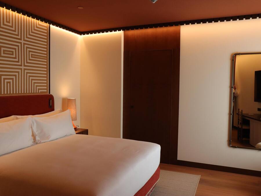A neatly made bed with white linens, a wooden headboard, a wall-mounted mirror, and a door with a wooden panel.