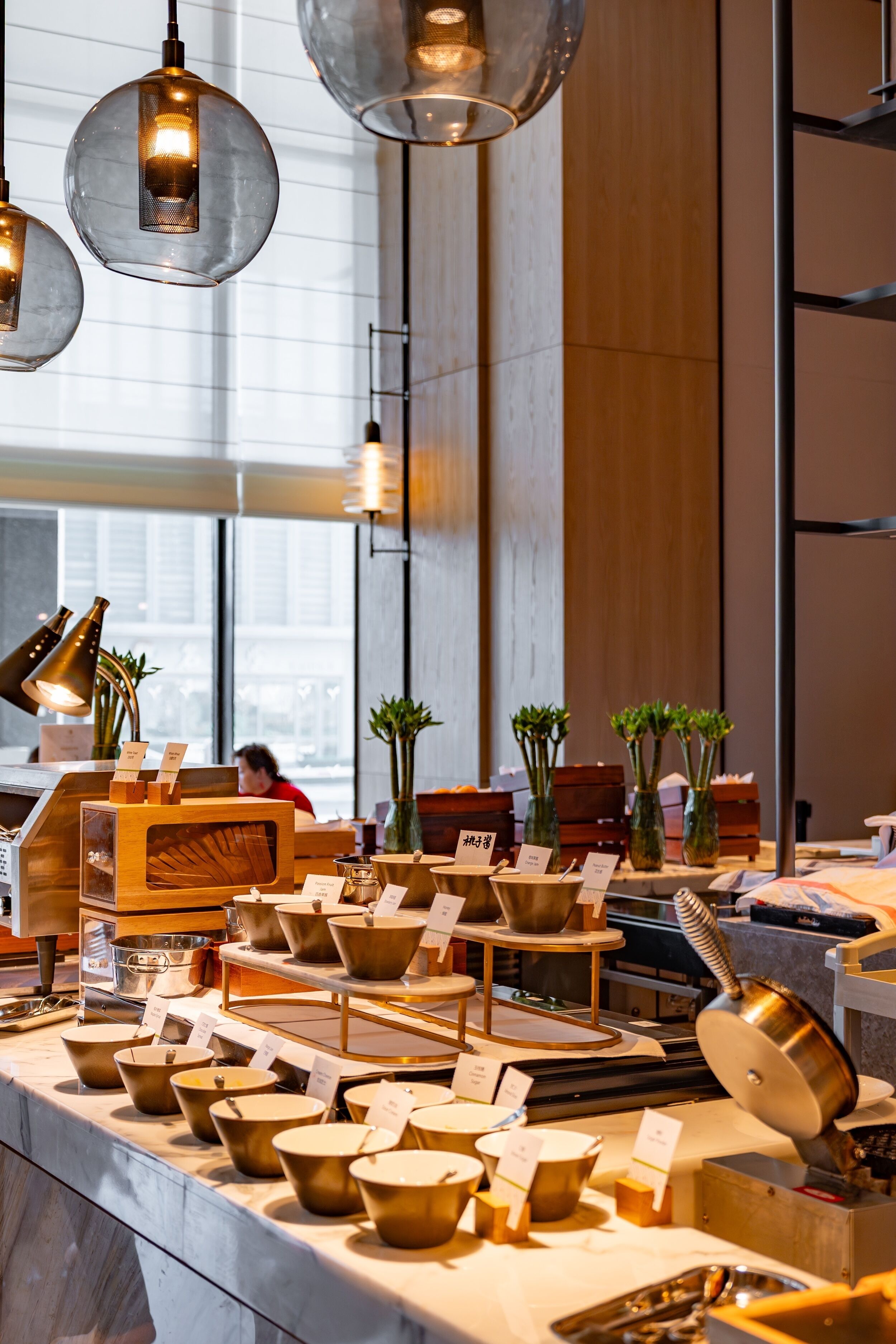A modern interior with a long counter displaying various bowls and utensils, overhead hanging lights, and a large window providing natural light.