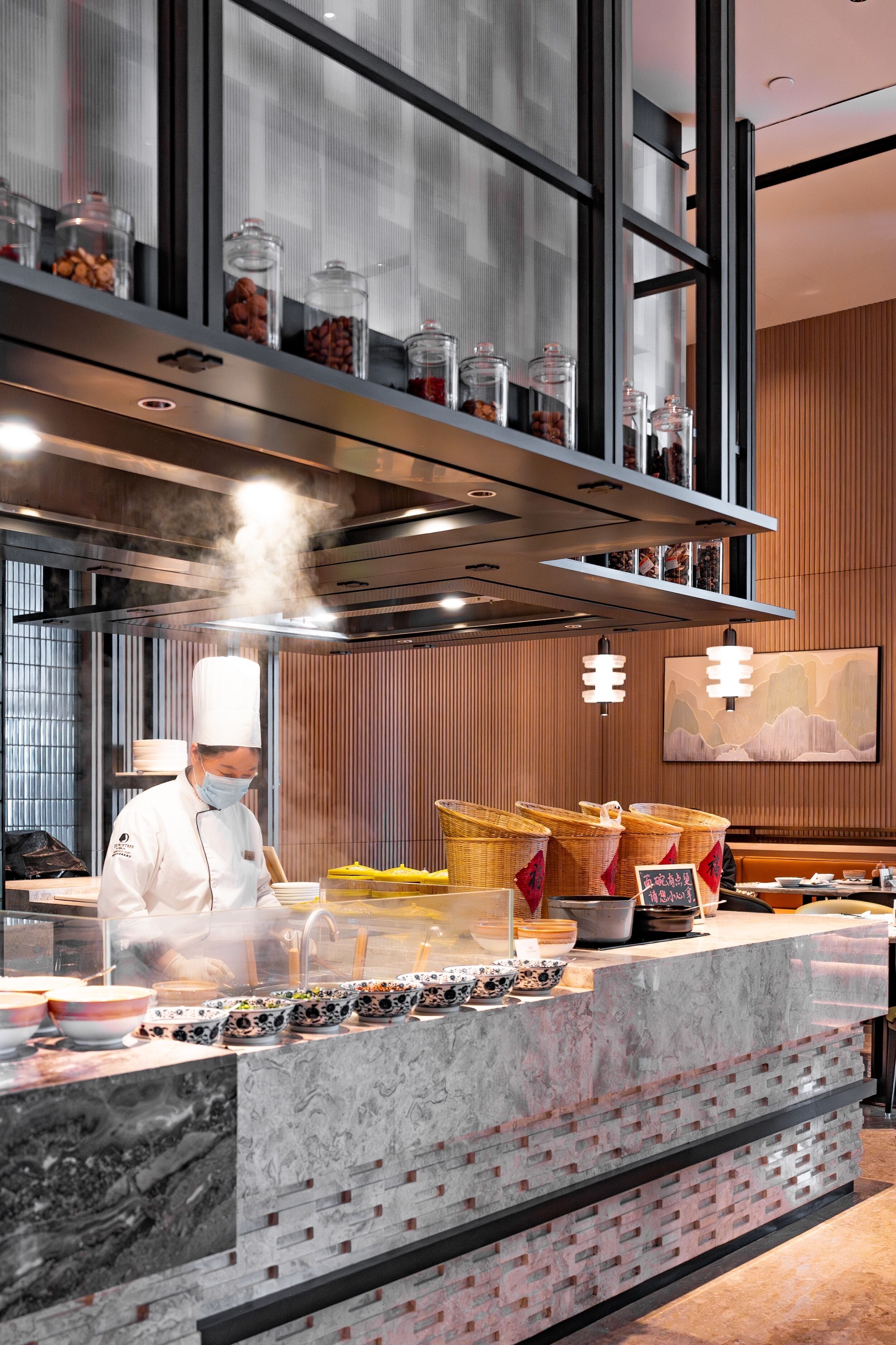 A modern kitchen with a chef preparing food, a marble countertop, and a high ceiling with a ventilation system.