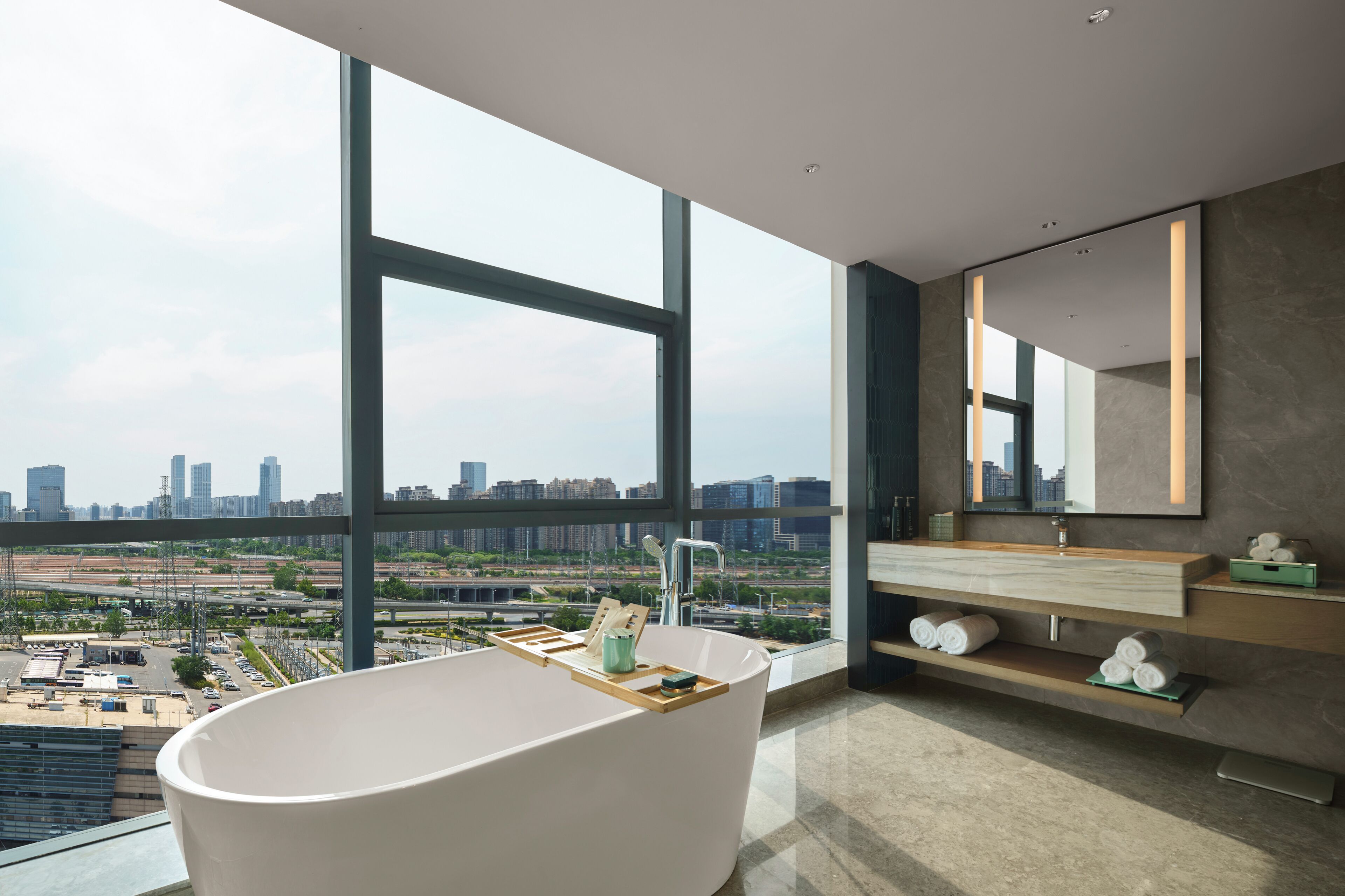 Un baño moderno con una bañera independiente, un espejo grande y vistas a un paisaje urbano a través de amplias ventanas.
