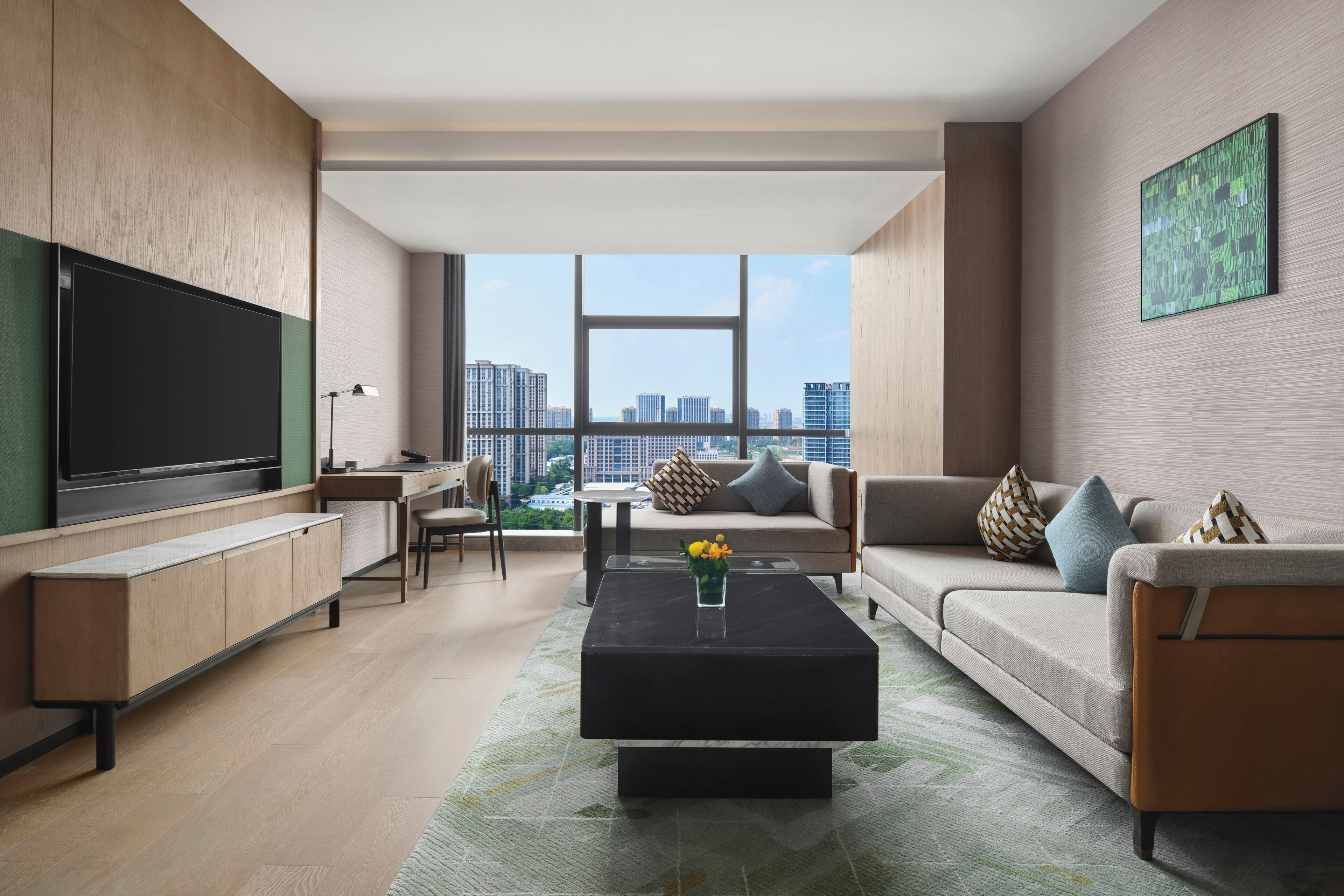 A modern living room with a flat-screen TV, a sofa, a coffee table, and a view of the cityscape through large windows.