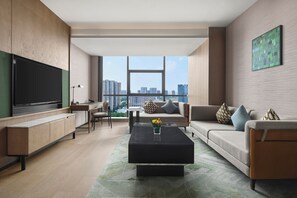 Un salón moderno con televisor de pantalla plana, un sofá, una mesa de centro y vistas a la ciudad a través de amplios ventanales.