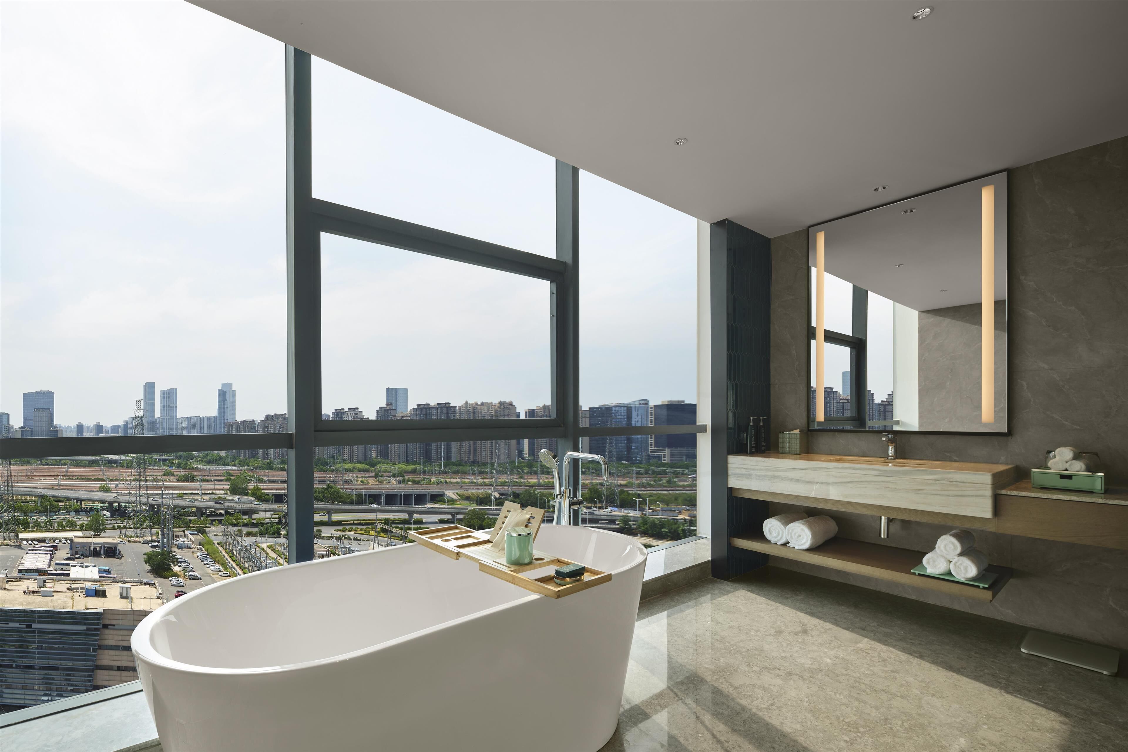 A modern bathroom with a freestanding bathtub, a large mirror, and a view of a cityscape through large windows.