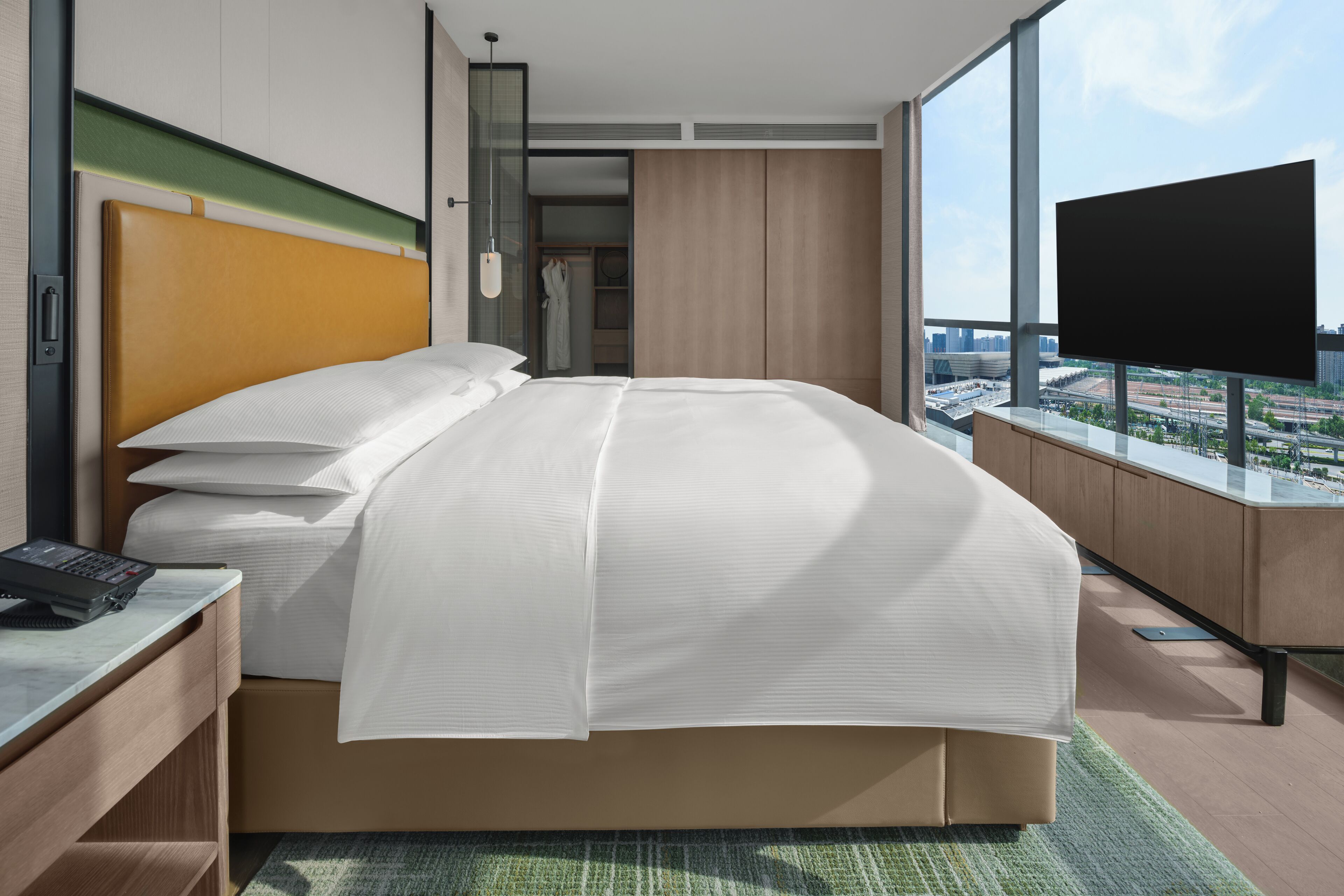 A modern hotel room with a large bed, a desk with a phone, a TV, and a view of the city through large windows.