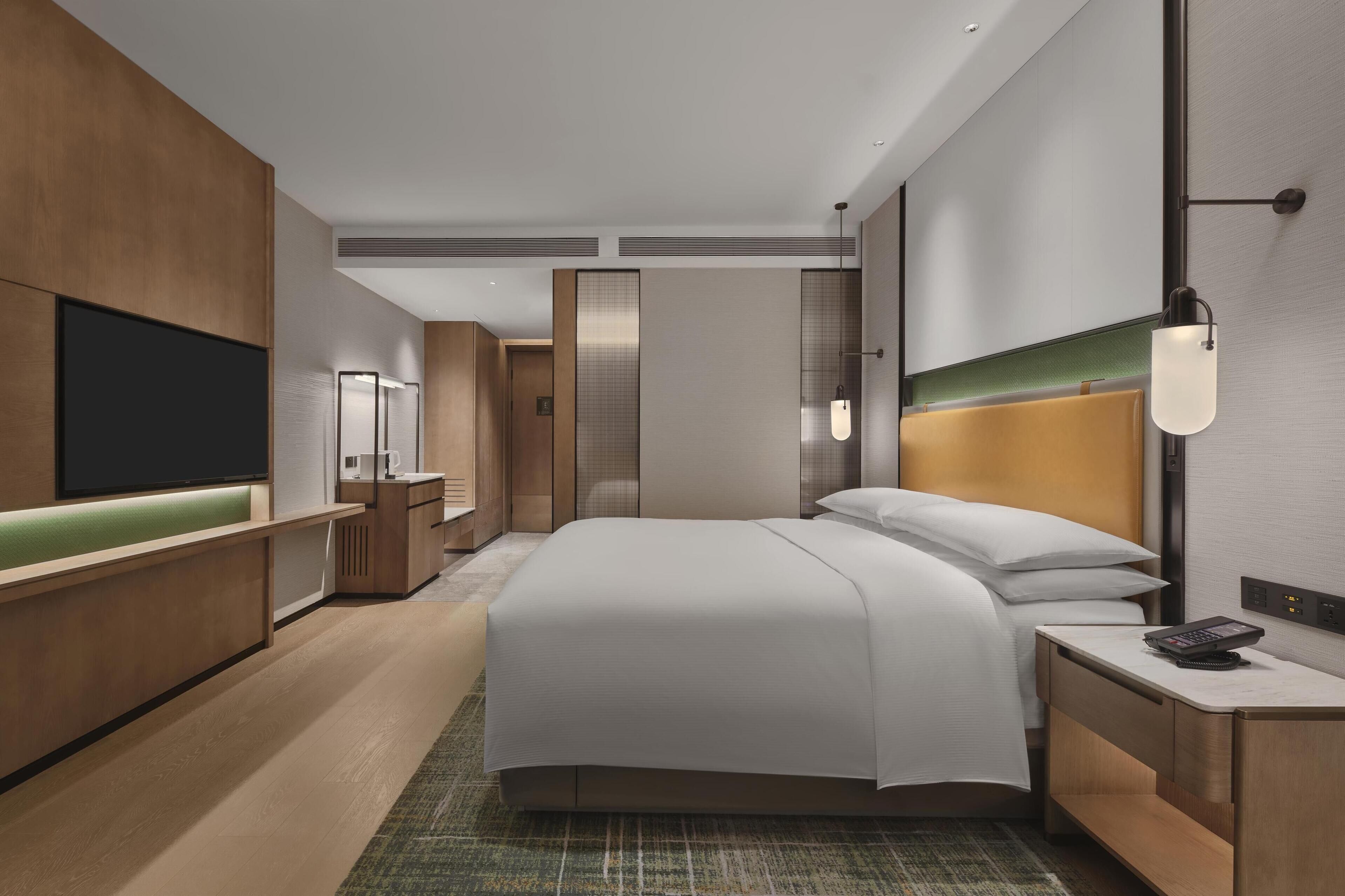 A modern hotel room with a large bed, a bedside table with a phone, a flat-screen TV mounted on the wall, and a separate seating area with a bench and a small table.