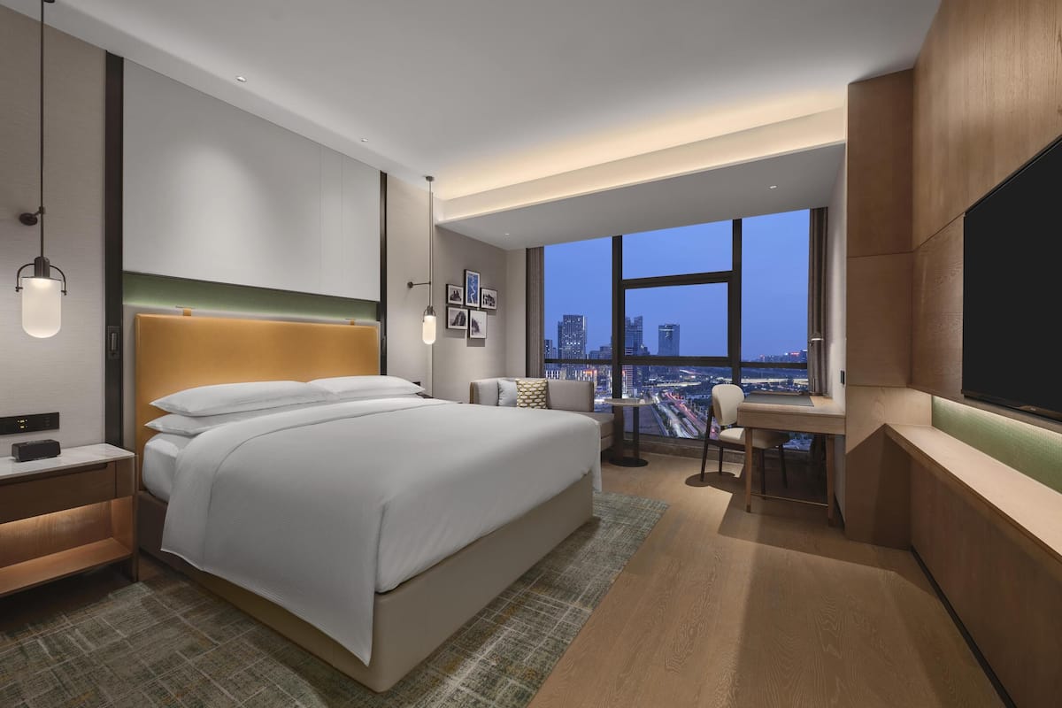 A modern hotel room with a large bed, a desk, and a view of the cityscape.