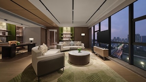 Una sala moderna con sofá, mesa de centro y televisor. Hay un escritorio con silla y una estantería. La habitación tiene ventanas amplias con vistas a la ciudad.