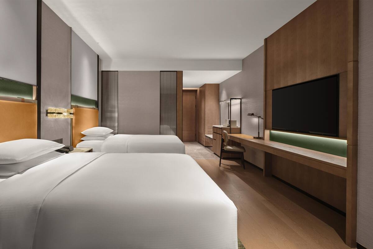 A modern hotel room with two beds, a desk, a chair, a TV, and a large mirror.