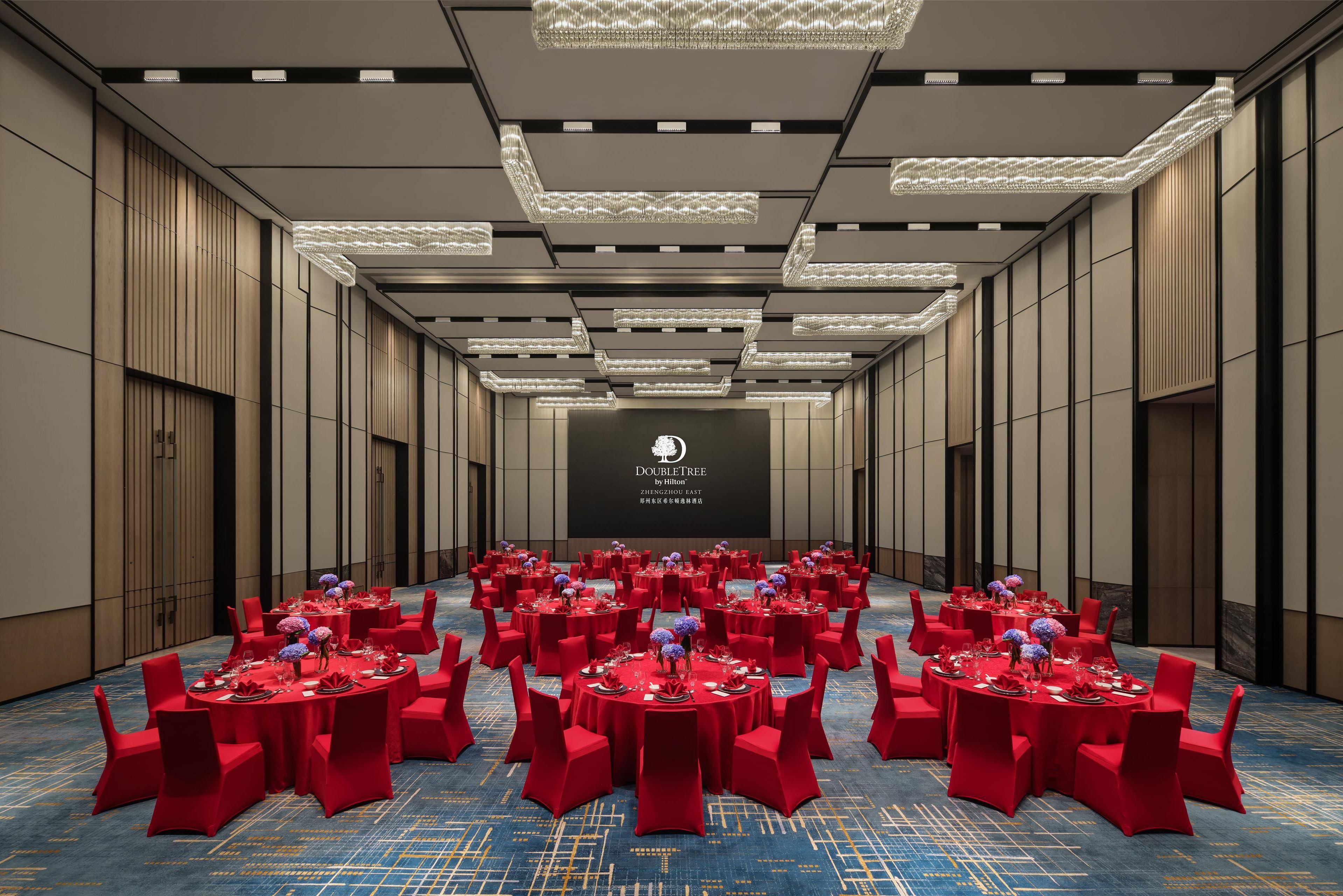 A conference room with multiple tables set for a formal event, featuring red chairs and tables, a large screen in the background, and a modern chandelier.