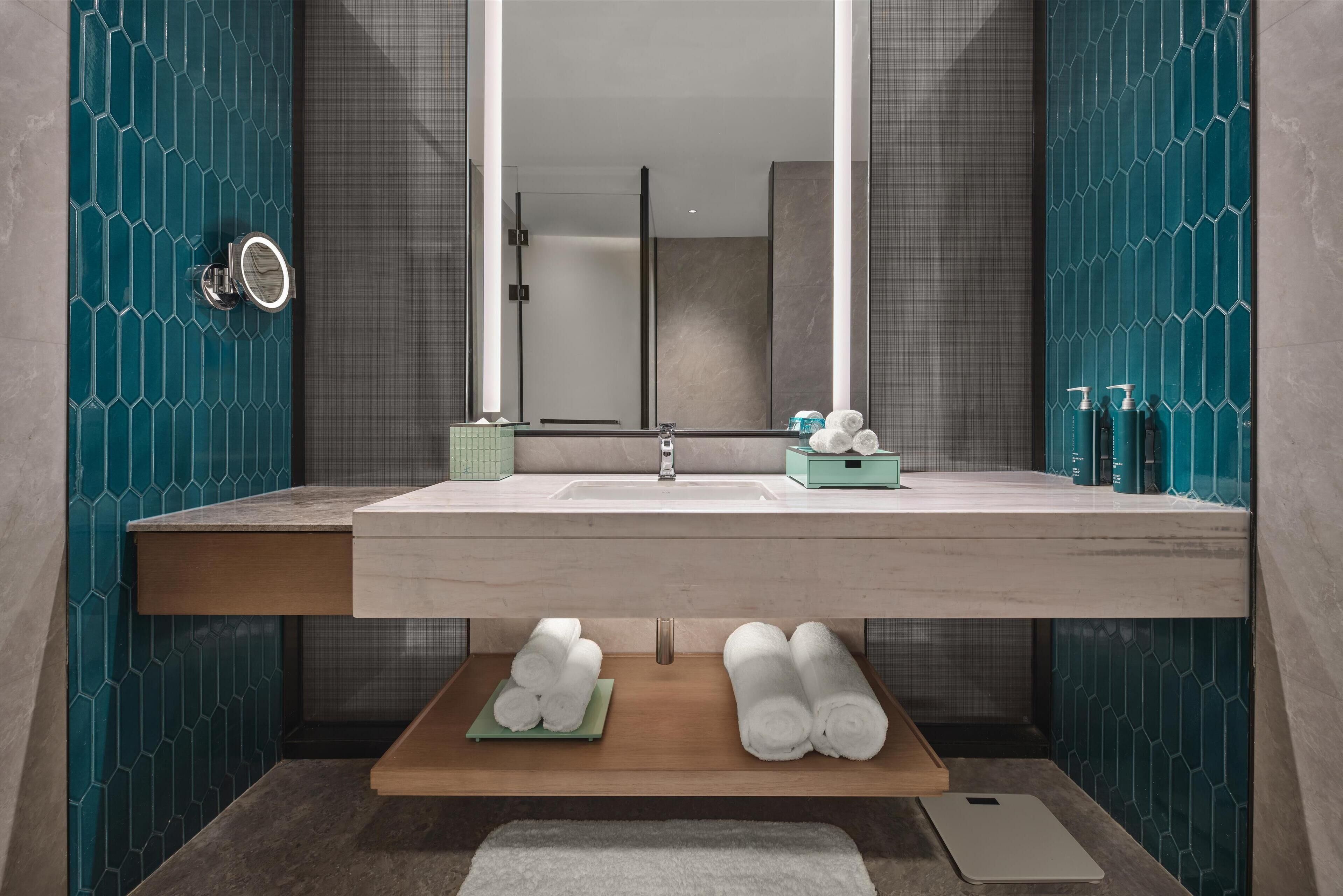 A modern bathroom with a large mirror, a sink, and a shower area. There are towels and toiletries on the counter.