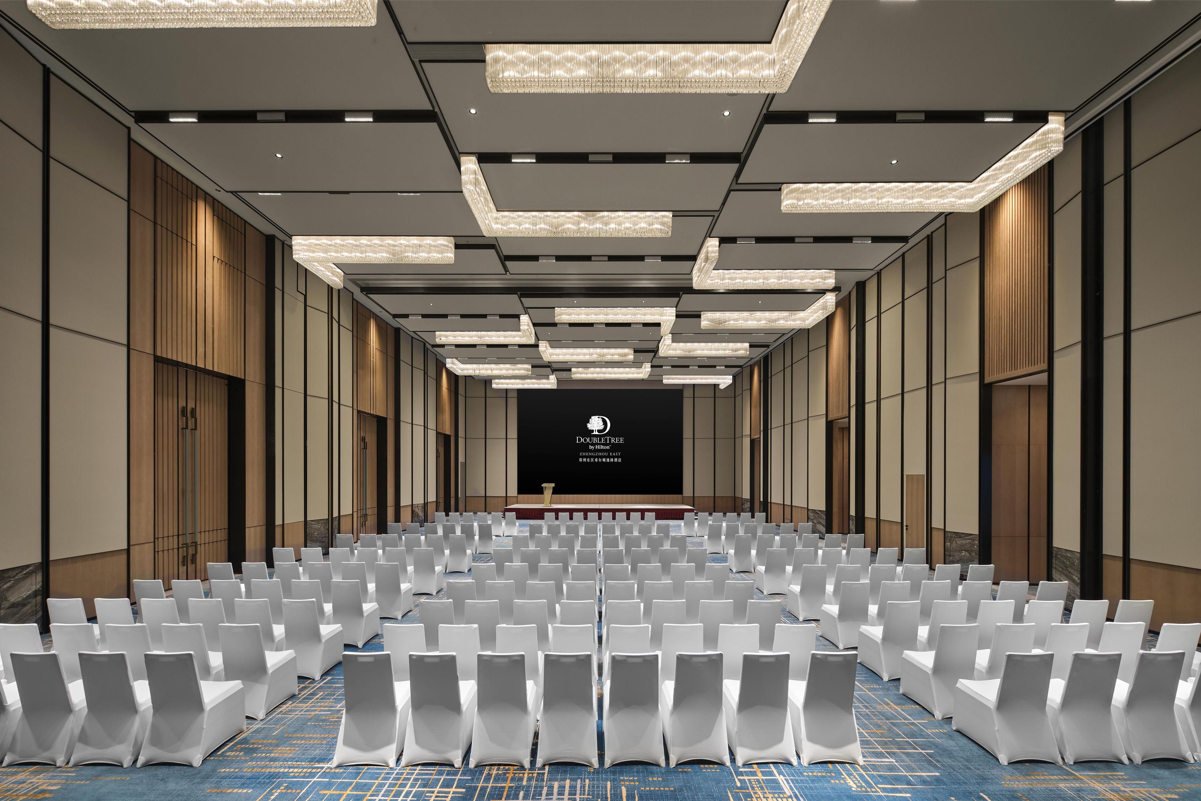 A conference room with rows of white chairs, a stage with a podium, and a large screen displaying text.
