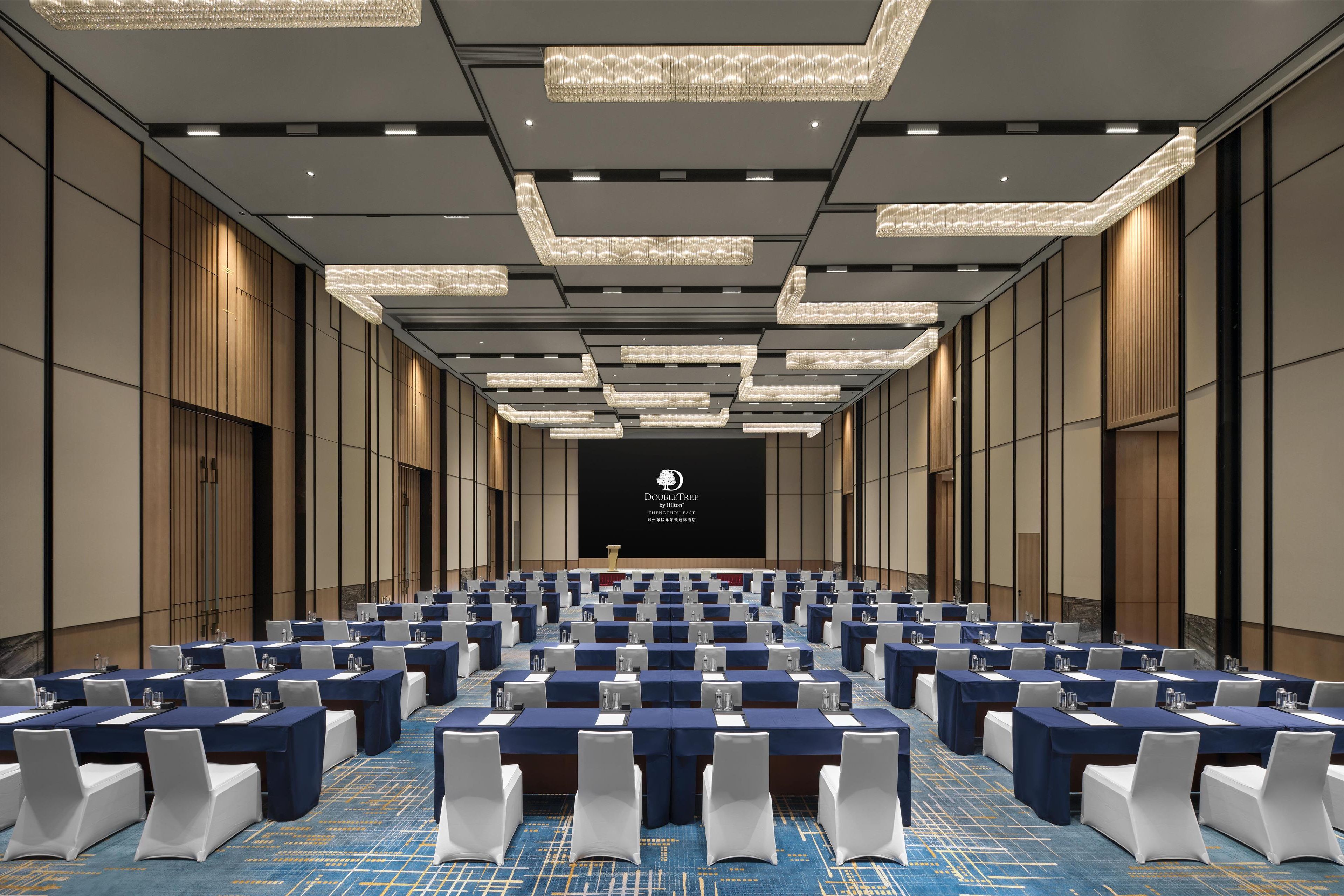 Een congreszaal met rijen tafels, witte stoelen en een groot scherm aan de voorkant.