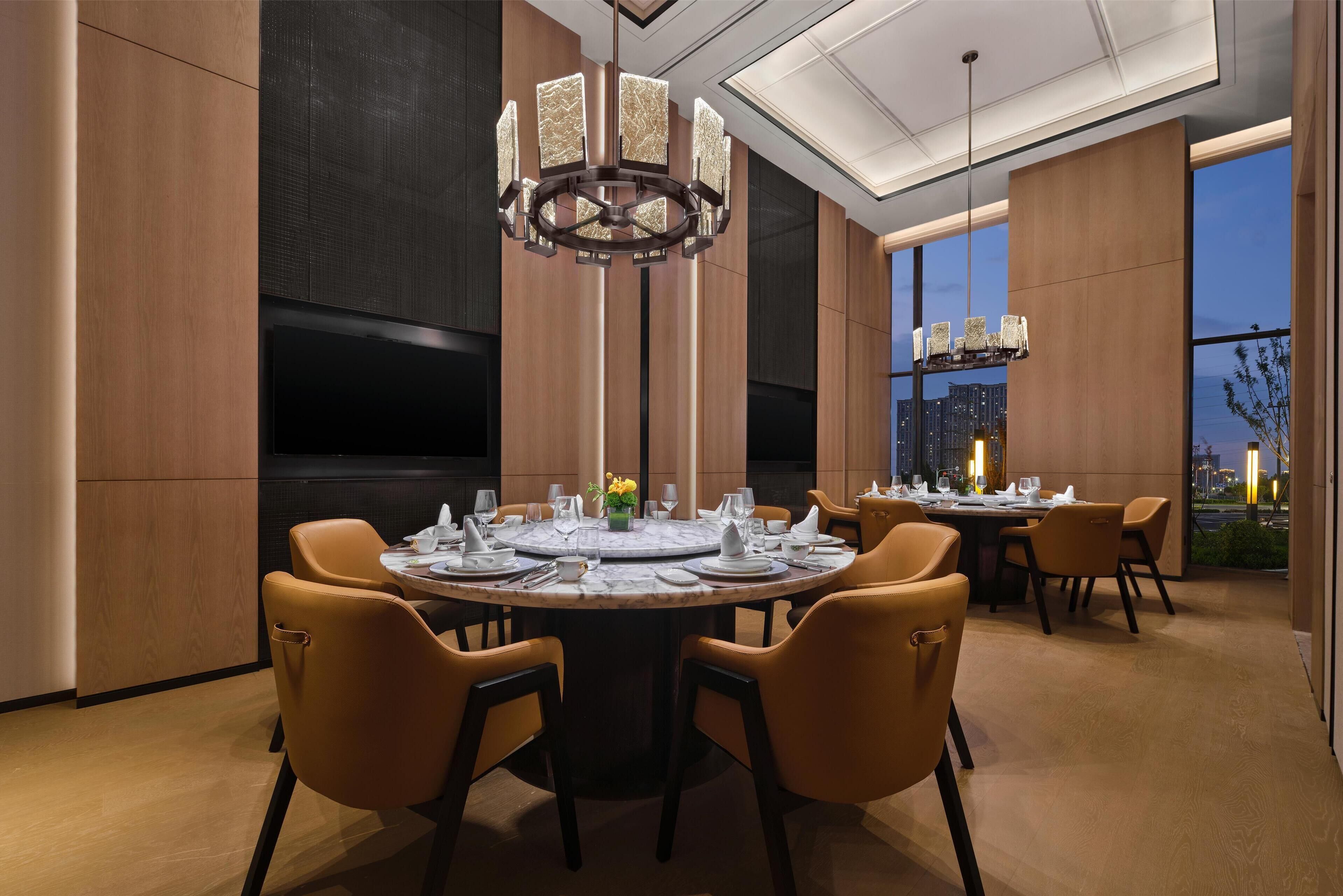 Een moderne eetruimte met een ronde tafel voor vier personen, omringd door gele stoelen. Een grote kroonluchter hangt aan het plafond, en de kamer heeft houten lambrisering en een groot raam met uitzicht op de stad.