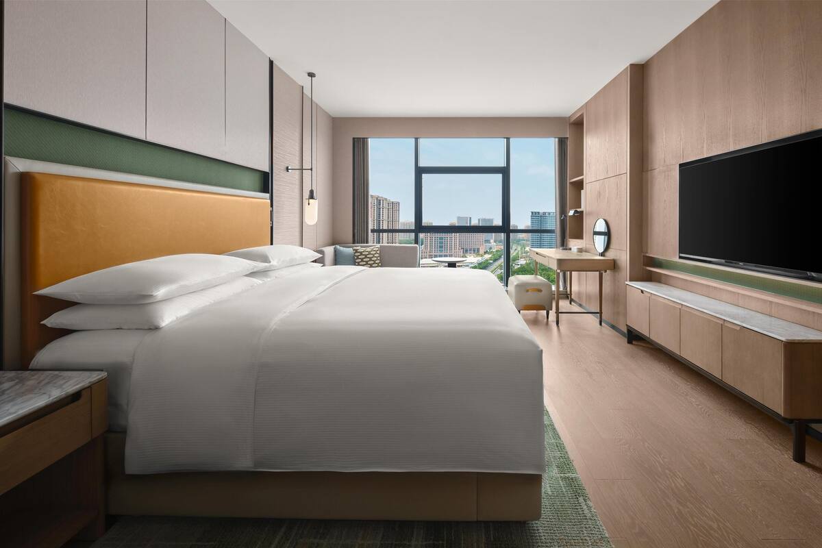 A modern hotel room with a large bed, a TV, a desk, and a view of the city.
