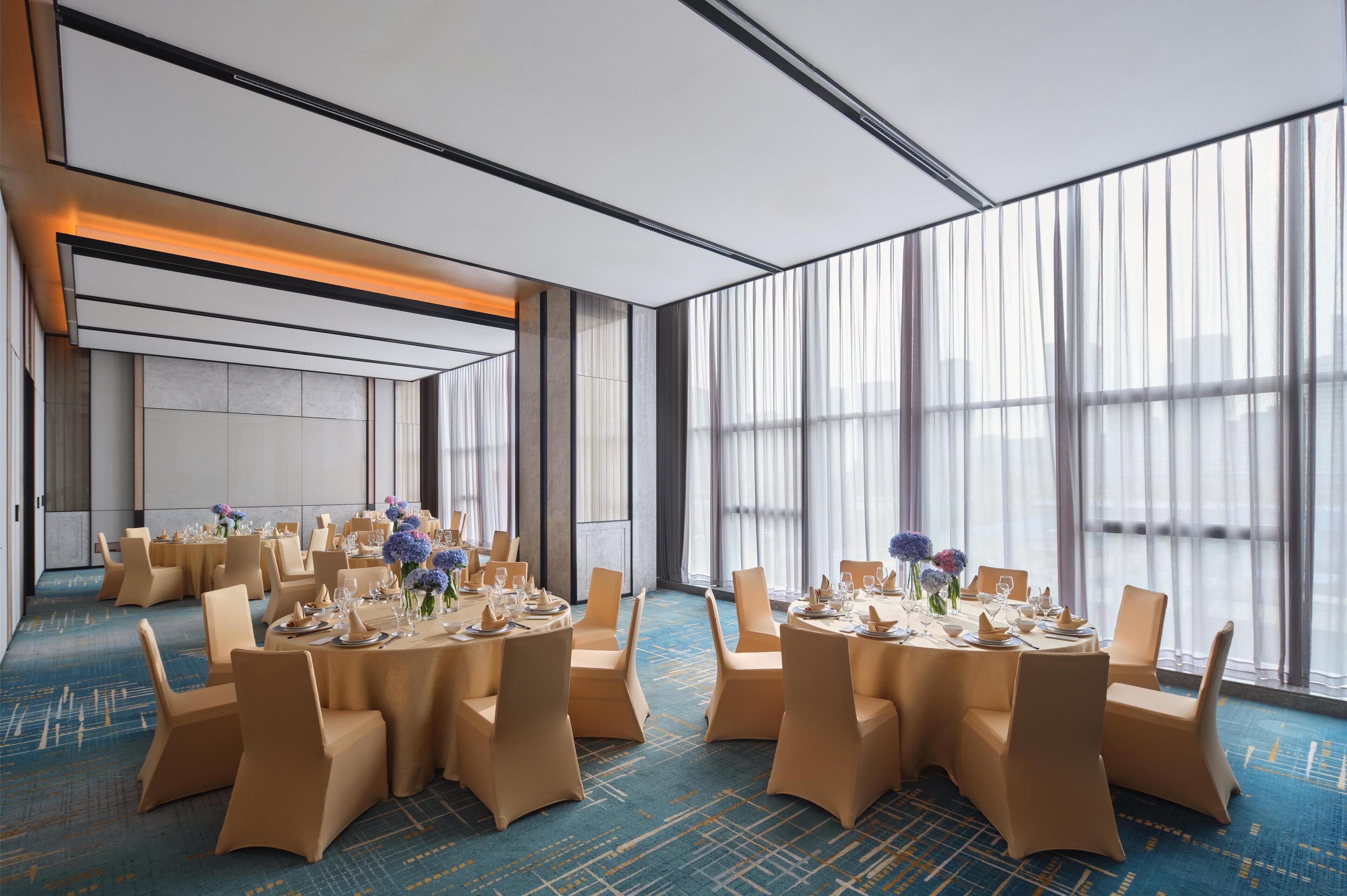 Un salón de conferencias amplio con mesas redondas, sillas color beige y grandes ventanales con vistas.
