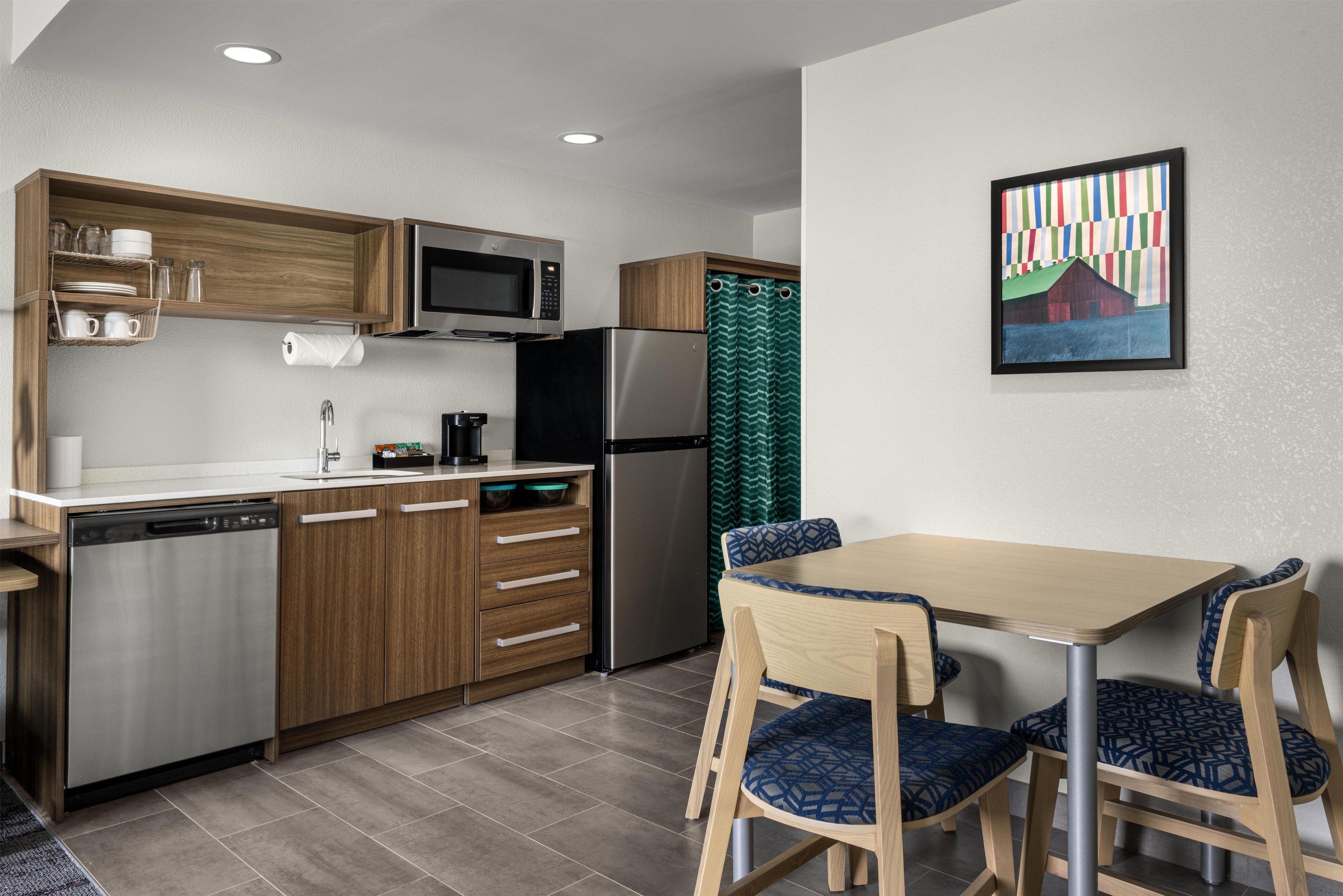 A modern kitchen with wooden cabinets, a stainless steel refrigerator, a microwave, and a dishwasher. There is a dining area with a wooden table and blue patterned chairs.