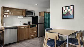 A modern kitchen with wooden cabinets, a stainless steel refrigerator, a microwave, and a dishwasher. There is a dining area with a wooden table and blue patterned chairs.