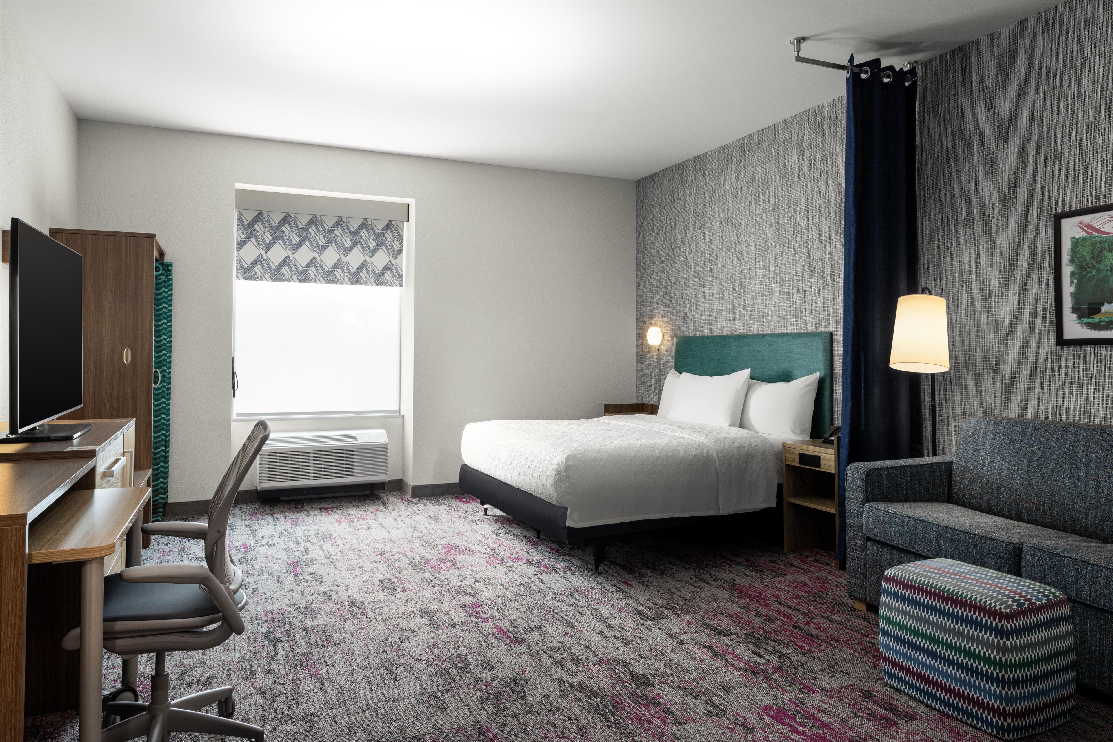 A hotel room with a bed, a desk, a chair, a TV, a window, and a painting on the wall.