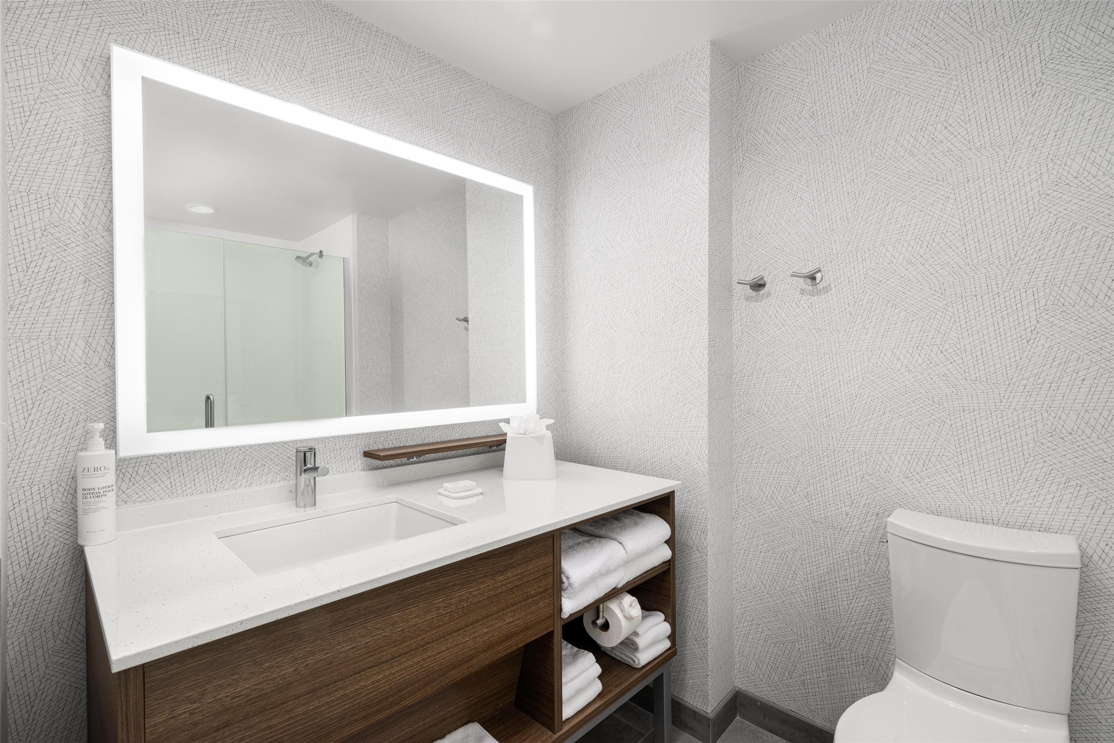 Un baño moderno con un espejo grande, lavamanos blanco, tocador de madera y un inodoro.