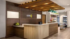 Ein Hotel-Rezeptionsbereich mit einem Holzbüro, einem Schild mit der Aufschrift 'HOME 2 Suites by Hilton' und einer Snackbar im Hintergrund.
