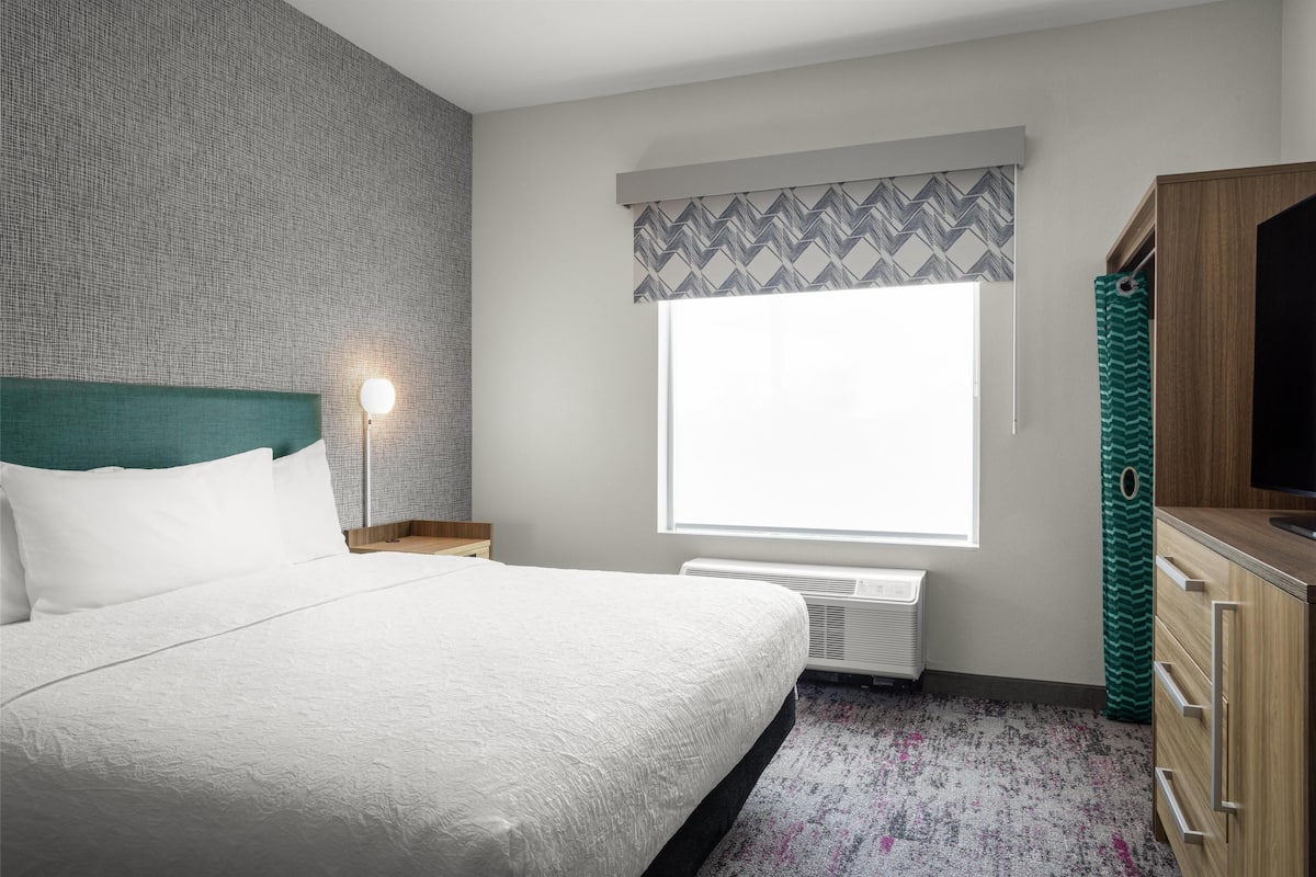 A hotel room with a large bed, a nightstand with a lamp, a window with patterned curtains, a radiator, and a wooden wardrobe.