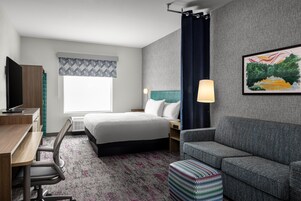 Una habitación de hotel con cama, escritorio con televisor, sofá, alfombra estampada y un cuadro en la pared.