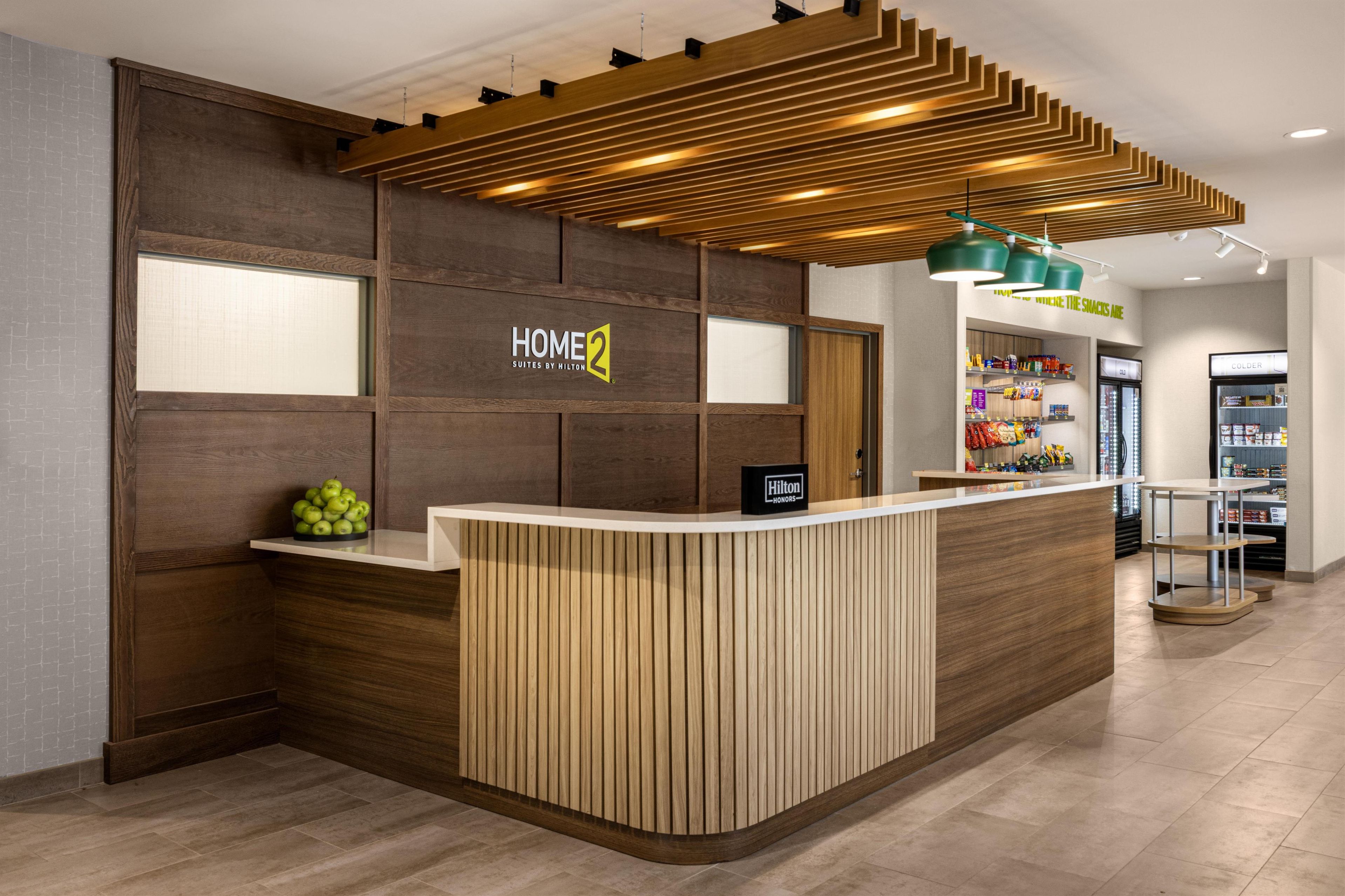 Zona de recepción de un hotel con un mostrador de madera, un cartel que dice "HOME 2 Suites by Hilton" y una barra de aperitivos al fondo.