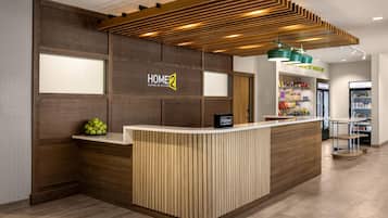 Zona de recepción de un hotel con un mostrador de madera, un cartel que dice "HOME 2 Suites by Hilton" y una barra de aperitivos al fondo.
