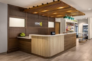 Zona de recepción de un hotel con un mostrador de madera, un cartel que dice "HOME 2 Suites by Hilton" y una barra de aperitivos al fondo.