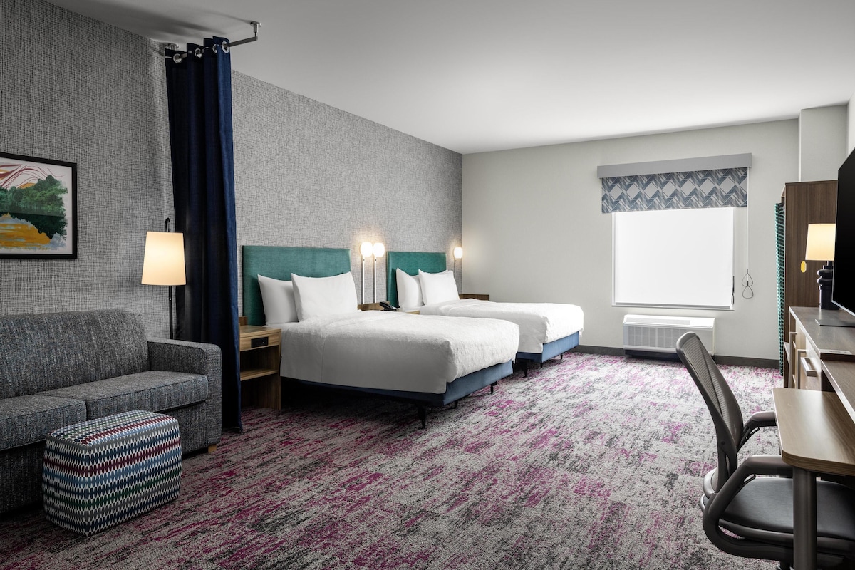 Quarto de hotel com parede cinza texturizada, uma janela grande com cortina estampada, sofá cinza, duas camas com roupa de cama branca, um pequeno banquinho e uma escrivaninha com cadeira.