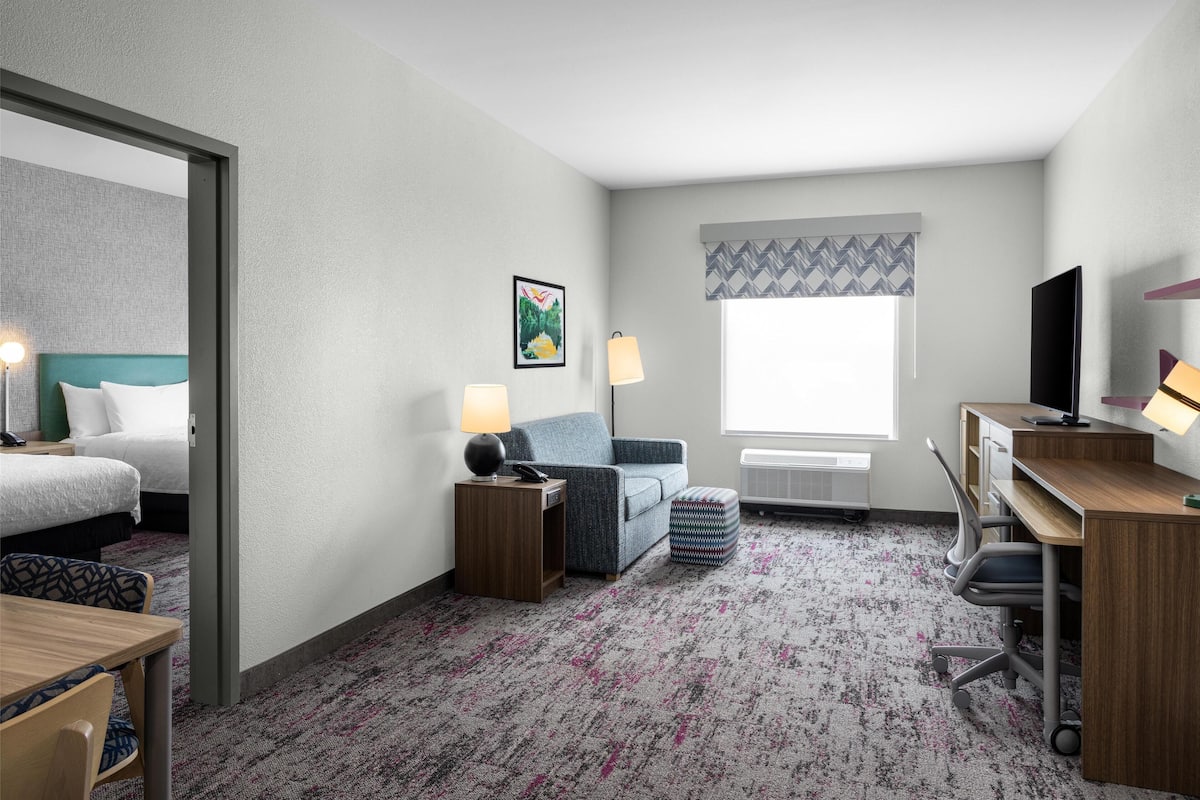 A hotel room with a bed, a desk, a chair, a sofa, a lamp, a TV, and a window with a patterned curtain.