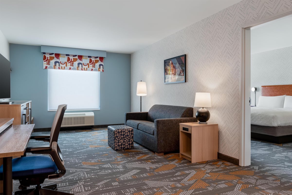 Habitación de hotel con escritorio, silla, sofá, reposapiés, dos lámparas de noche, cama y ventana grande.