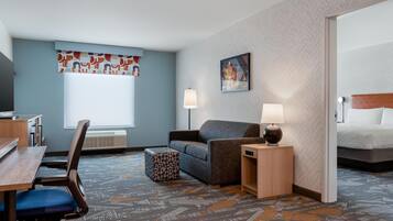 A hotel room with a desk, chair, sofa, ottoman, two bedside lamps, a bed, and a large window.