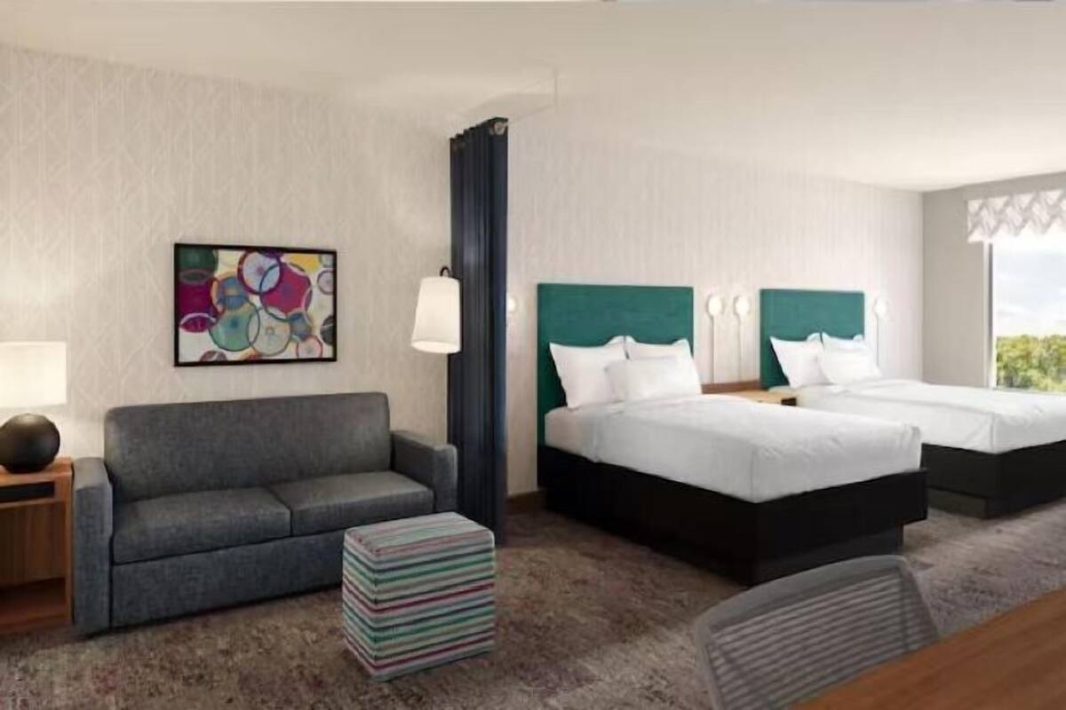 Habitación de hotel con sofá, dos camas con ropa blanca, un cuadro abstracto de colores y un ventanal grande.