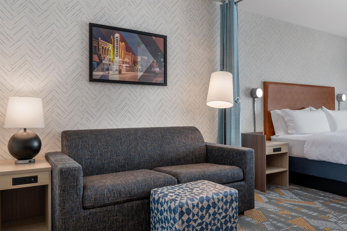 Ein Hotelzimmer mit Sofa, Hocker, Nachttisch, Lampe, Beistelltisch und einem Bett mit Kopfteil.